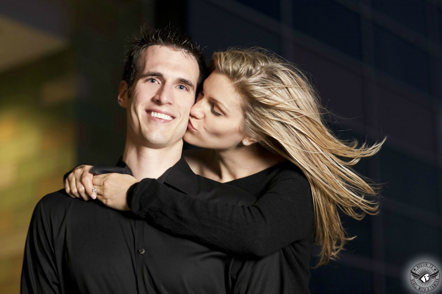 Happy dark spiky haired guy in a black button up dress shirt is hugged around the neck and kissed on the cheek by a hot windswept blond lady wearing a black crew sweater on the stairs at the Long Center in this happy engagement portrait in Downtown Austin, Texas. 